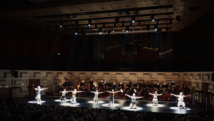 In de sfeervol uitgelichte Grote Zaal van de Doelen knielen acht dansers van Conny Janssen Danst in een cirkel van licht, met gespreide armen, tijdens de uitvoering van Górecki’s Symphony of Sorrowful Songs. Op de achtergrond speelt Sinfonia Rotterdam, met centraal de sopraan  Ewa Tracz en dirigent Conrad van Alphen zichtbaar voor het orgel.