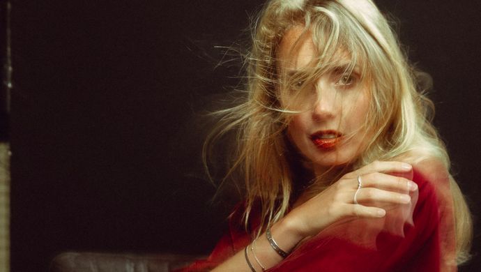 Woman with long blonde hair wearing a red velvet dress sits on a dark leather couch, gazing at the camera with a confident, slightly mysterious expression.