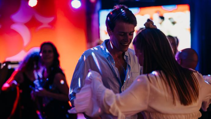 A man and a woman are dancing together during Disco Royale at de Doelen Studio. They are having fun on the dance floor, surrounded by colorful lighting and other partygoers in the background. The atmosphere is festive and intimate.