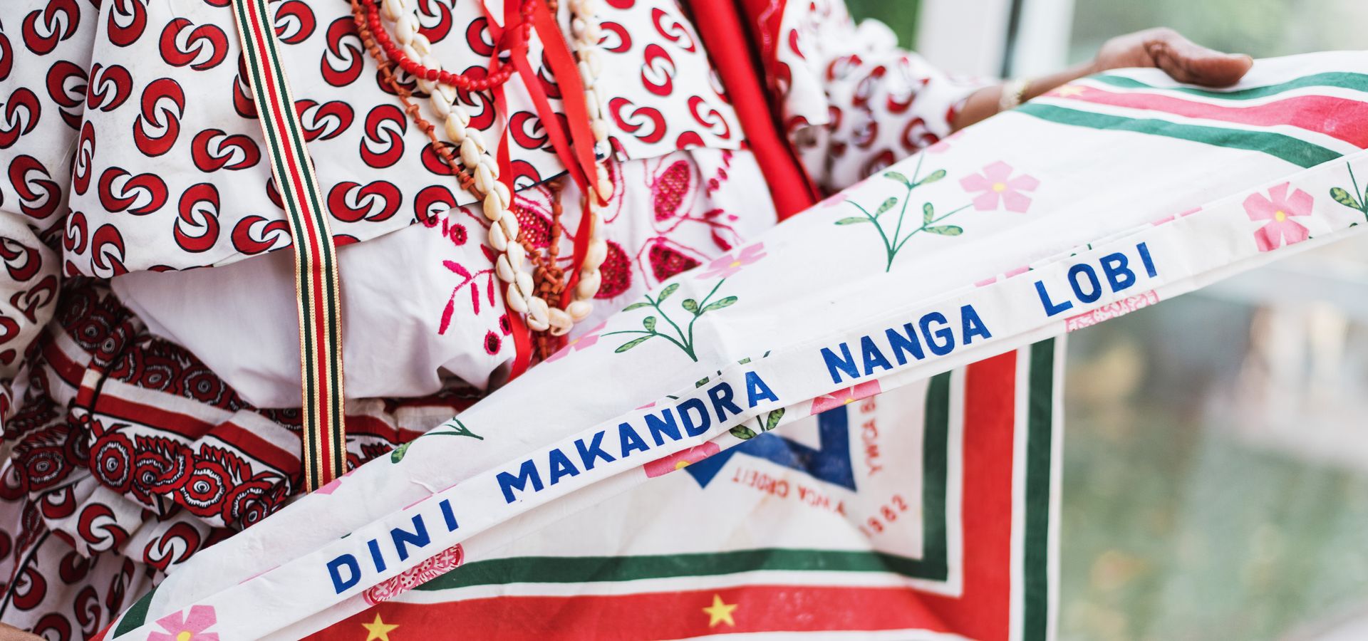 Persoon in traditionele kleding houdt een kleurrijke driehoekige sjaal vast waarop de tekst staat: "Ratu Makandre Tangi Libi".