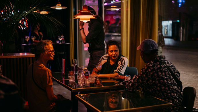 Three people sit at a table engaged in conversation during Disco Royale at de Doelen. They are in a warmly lit space with glasses on the table and hanging lamps above. Through the window, the dance floor with purple lighting is visible in the background.
