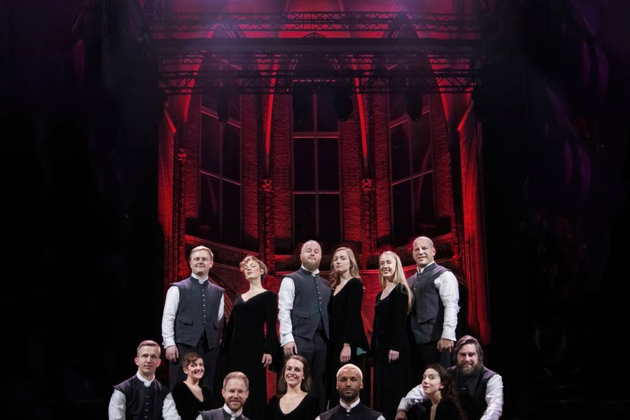 Groepsfoto van een koor van twaalf personen in formele zwarte kleding met witte accenten, poserend op een trap met een dramatisch rood verlicht decor op de achtergrond.