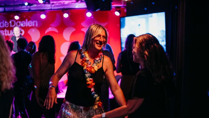 A festive atmosphere during the Disco Royale event at Studio 1 of de Doelen. In the foreground, a smiling woman is dancing, wearing a black top, a silver glitter skirt, and a colorful flower lei. In the background, other visitors are dancing under purple and red stage lights, with the de Doelen Studio logo visible on the wall.