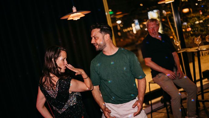 A man and a woman dance cheerfully together during Disco Royale at de Doelen. They laugh and make eye contact, while another guest relaxes in the background in a warmly lit space with a view outside.