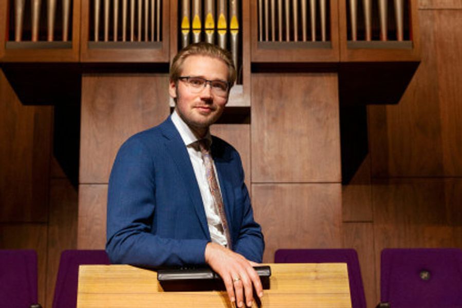 Een man in een blauw colbert met zwarte vlinderdas staat in een kerk of concertzaal voor een houten orgel. Hij draagt een bril en kijkt recht in de camera, met één hand leunend op een orgelbank. De ruimte heeft een klassieke uitstraling met paarse stoelen op de achtergrond.