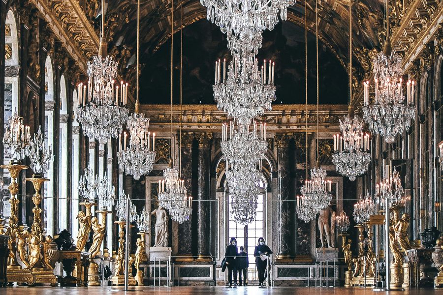 Rijkelijk versierde spiegelzaal met kristallen kroonluchters, gouden ornamenten en marmeren zuilen, vermoedelijk in het Paleis van Versailles.