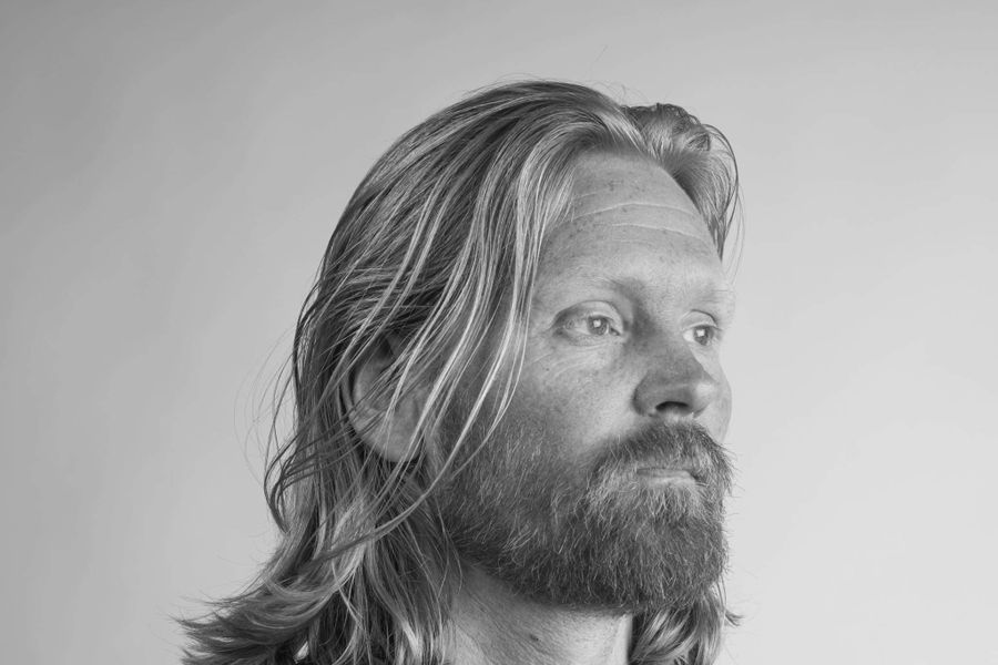 Zwart-wit portret van een man met halflang haar en baard, die licht van de camera wegkijkt met een rustige uitdrukking.