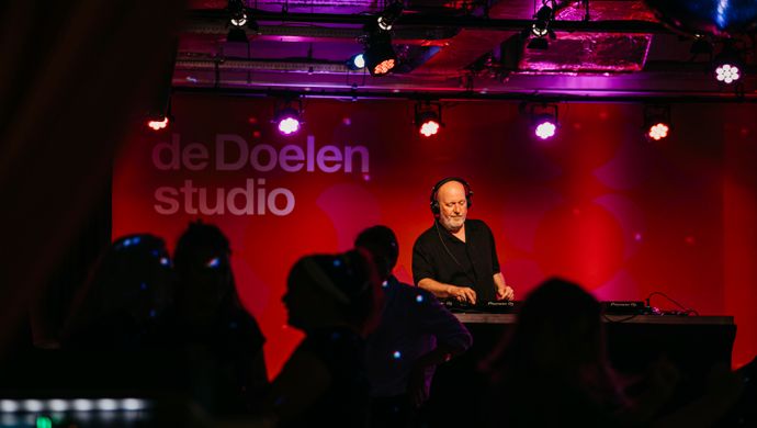 A DJ is playing music during the Disco Royale event at Studio 1 of de Doelen. In the foreground, silhouettes of dancing guests are visible in the dark, while the DJ stands on a stage with a red backdrop displaying the de Doelen Studio logo. Purple and red stage lighting enhances the festive atmosphere of the space.