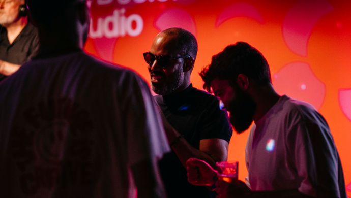 Two men are standing close together in conversation during the Disco Royale event. In the background, a red-lit wall adds to the vibrant atmosphere.