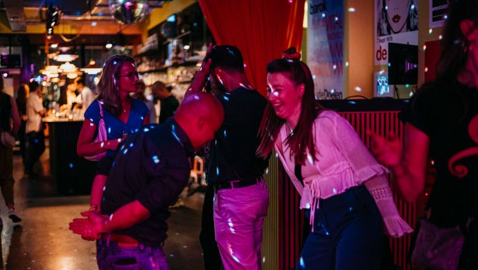 Partygoers dance joyfully during Disco Royale at de Doelen Studio. A man and woman laugh and move to the music, with colorful lighting and a bustling bar adding to the energetic atmosphere.