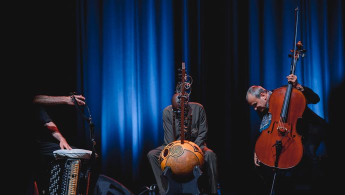 Vier muzikanten spelen in een intieme, blauw verlichte setting op accordeon, kora, cello en een klein snaarinstrument tijdens een sfeervol podiumoptreden.