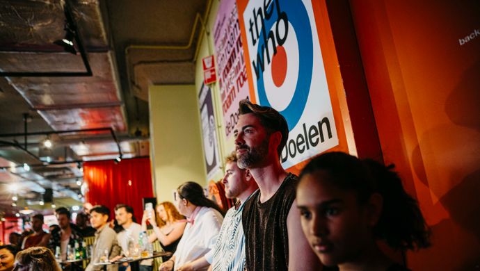 Bezoekers staan tegen een muur met kleurrijke posters terwijl ze naar de optredens op het podium kijken.