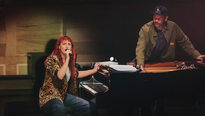 Wende sits at the piano and sings to the audience, with a man wearing a cap watching in the background.