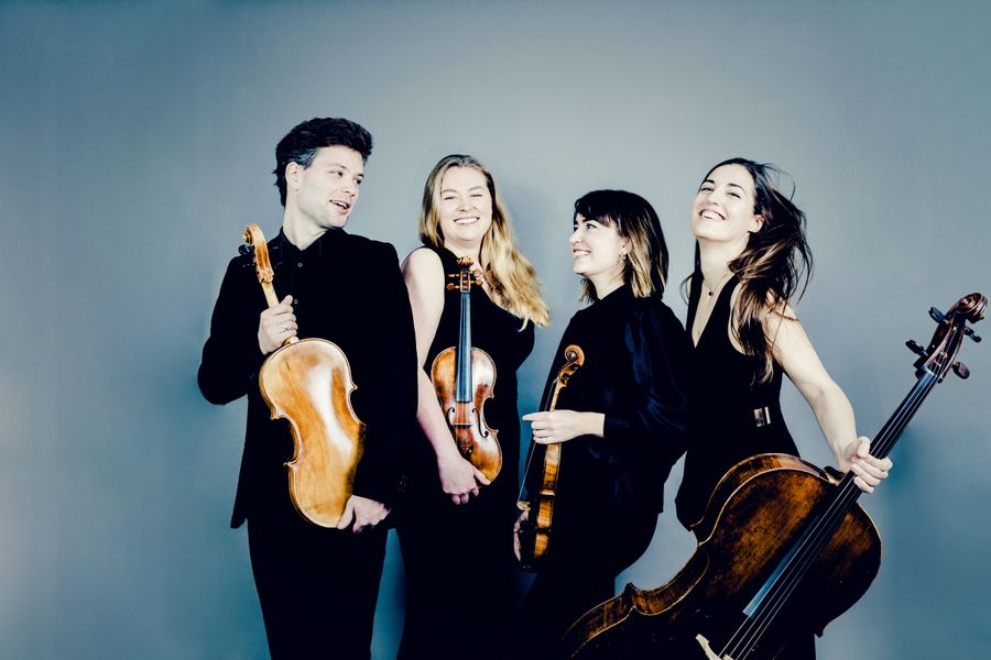 Vier klassieke musici poseren vrolijk samen voor een neutrale blauwe achtergrond. Twee vrouwen houden een viool vast, één een cello, en de man links een altviool. Ze lachen en maken oogcontact met elkaar.