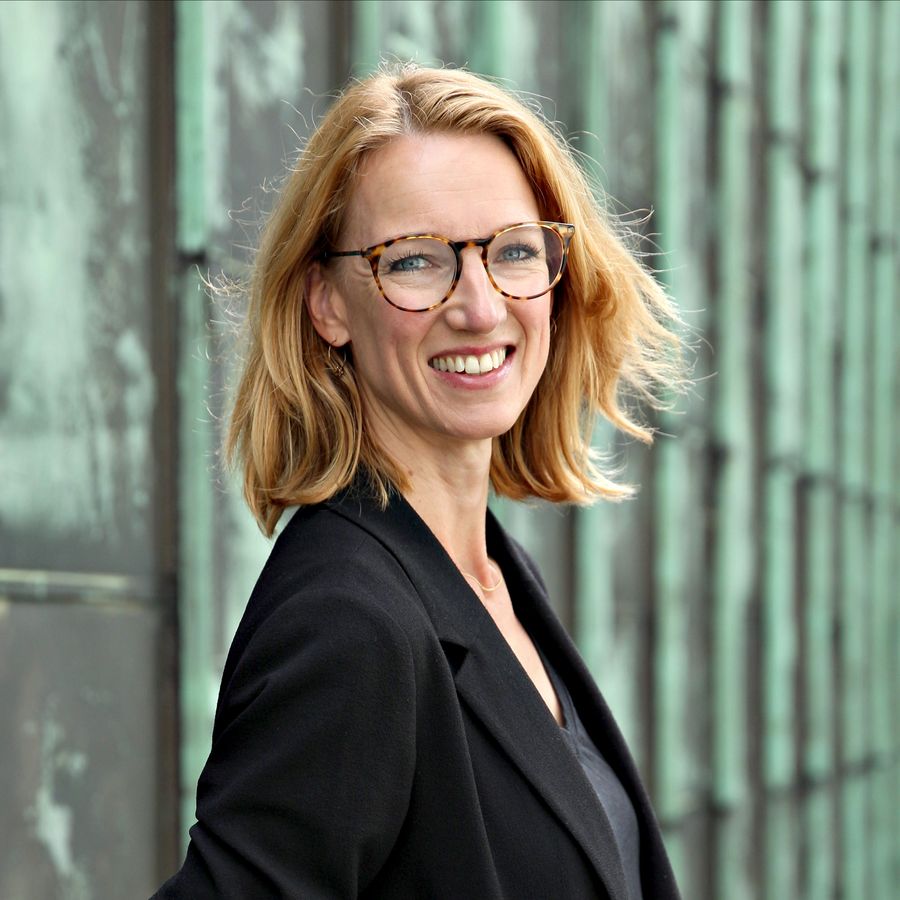 Vrouw met blond haar en bril glimlacht naar de camera, gekleed in een zwarte blazer, poserend buiten voor een verweerde metalen wand.