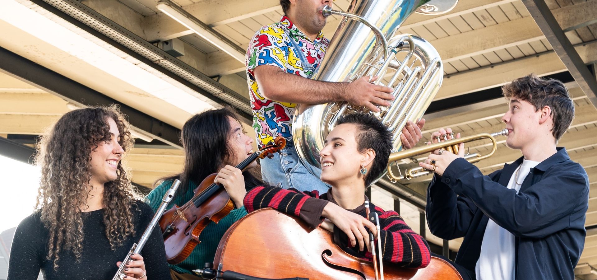 Vijf jonge muzikanten spelen klassieke instrumenten op een stedelijke buitenlocatie onder een viaduct. Ze bespelen onder andere viool, cello, klarinet en tuba, en hebben een energieke en vrolijke uitstraling.
