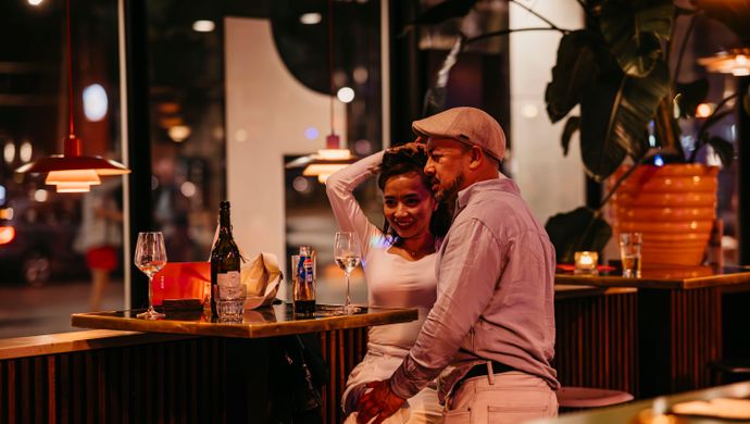 A couple sits close together at a table during Disco Royale at de Doelen. They smile and look at each other in a cozy bar setting with candlelight, bottles on the table, and the city visible through the windows in the background.