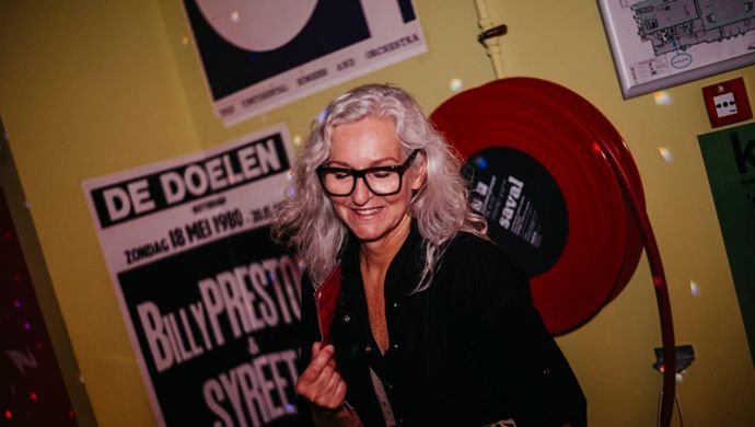 A smiling woman with long grey hair and glasses poses for the camera during Disco Royale at de Doelen. She stands in front of a retro-inspired wall decorated with colorful posters and a red fire hose reel.