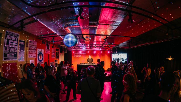 Overview of a packed dance floor during Disco Royale at de Doelen Studio. The venue is lit in red and purple, with a disco ball hanging from the ceiling and partygoers dancing in front of the DJ stage.
