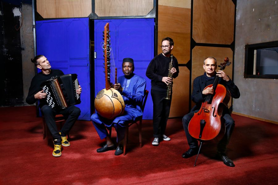 Vier muzikanten poseren met hun instrumenten — accordeon, kora, klarinet en cello — tegen een achtergrond van blauwe en houten panelen in een ruimte met rode vloerbedekking.