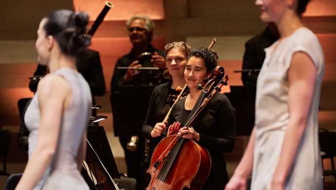 Een celliste van Sinfonia Rotterdam glimlacht terwijl ze haar instrument vasthoudt tijdens het slotapplaus in de Grote Zaal van de Doelen. Op de voorgrond lopen dansers van Conny Janssen Danst voorbij. De sfeer is warm en voldaan na afloop van de uitvoering van Górecki’s Symphony of Sorrowful Songs.