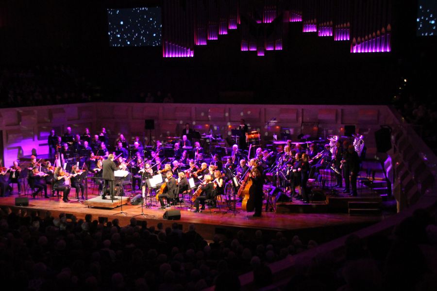 Orkest en koor voeren een concert uit op een podium met paarse sfeerverlichting. De zaal is gevuld met publiek en de musici zitten in een halve cirkel opgesteld.