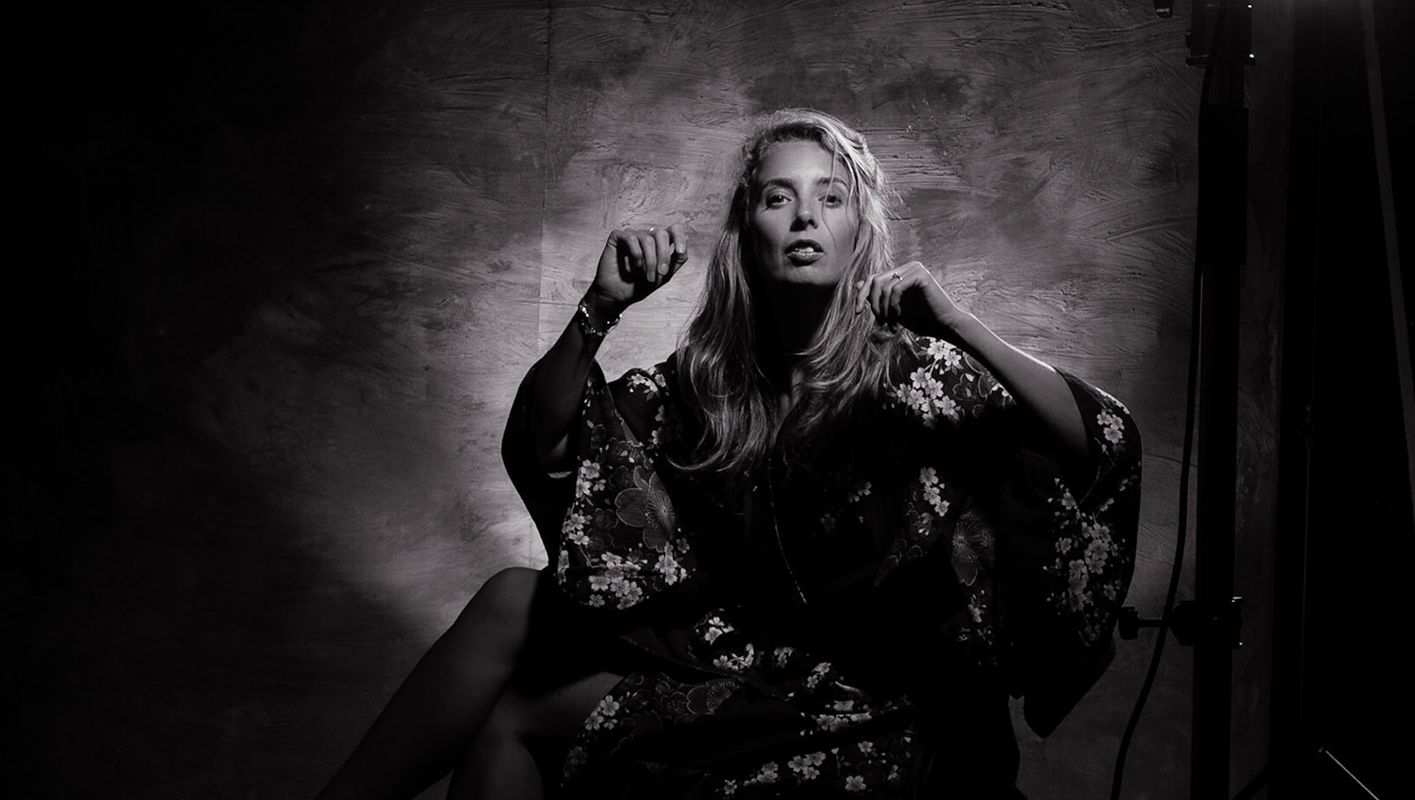 Black-and-white portrait of a woman with long hair in an expressive pose, wearing a voluminous floral-patterned garment. She is backlit, creating a halo-like glow around her.