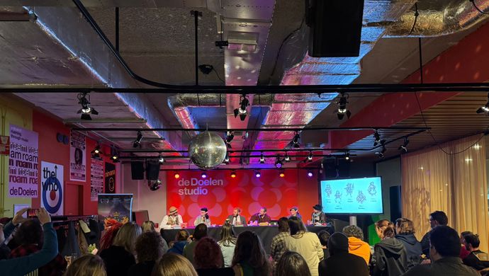 Publiek kijkt naar een paneldiscussie of presentatie in een moderne ruimte met rode achtergrond en paarse verlichting. Op het podium zitten sprekers aan een tafel en rechts is een scherm met projectie zichtbaar.