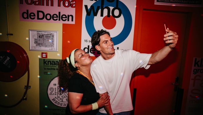 A man and a woman take a joyful selfie during Disco Royale at de Doelen. They stand in front of a colorful wall with posters from the de Doelen, while disco ball lights shimmer across the space.