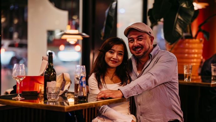 A smiling couple poses together at a standing table during Disco Royale at de Doelen. They sit close to each other with drinks and a bottle of wine on the table, surrounded by cozy lighting and inviting bar details.
