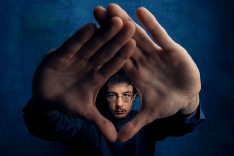 Portret van een man met donker haar tegen een diepe blauwe achtergrond. Hij kijkt serieus door een driehoek die hij met zijn handen vormt voor zijn gezicht, alsof hij door een lens kijkt.