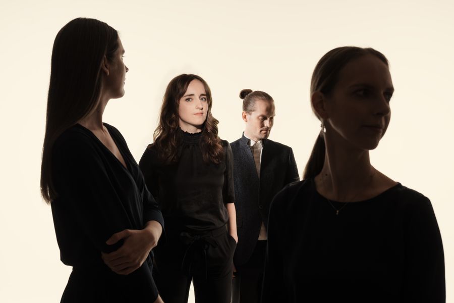 Vier vrouwen in zwarte kleding staan tegen een lichte achtergrond, deels in silhouet, met twee vrouwen scherp in beeld en twee in de schaduw op de voorgrond.