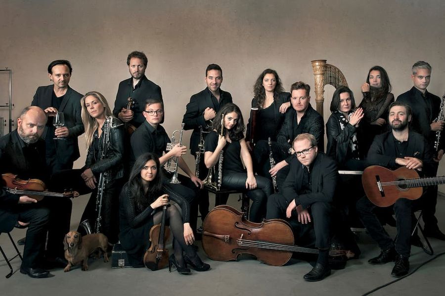 Groepsportret van een ensemble van zestien muzikanten met diverse instrumenten, zoals luit, viool, harp en fluit, geposeerd tegen een neutrale achtergrond in donkere kleding.