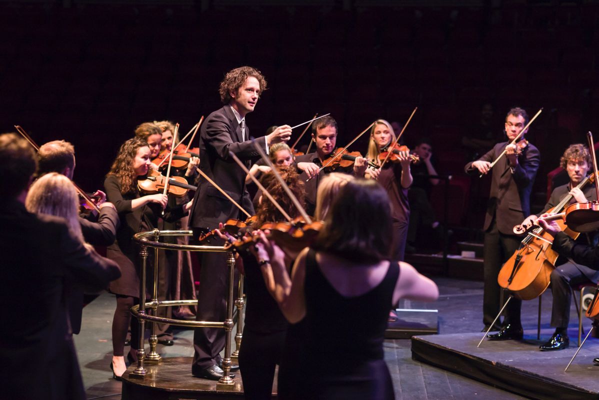 Dirigent leidt een strijkorkest op een podium; de musici spelen staand en in een halve cirkel, gekleed in concertkleding en intens geconcentreerd.
