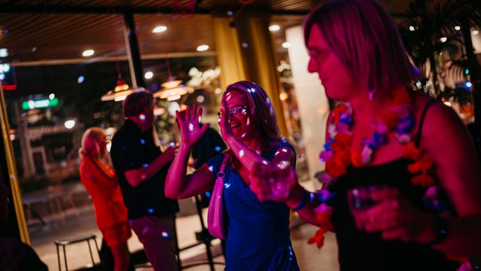 Two women dance and laugh together during Disco Royale at de Doelen, holding drinks in their hands. They are in a space with large windows and colorful light reflections, with other partygoers visible in the background.