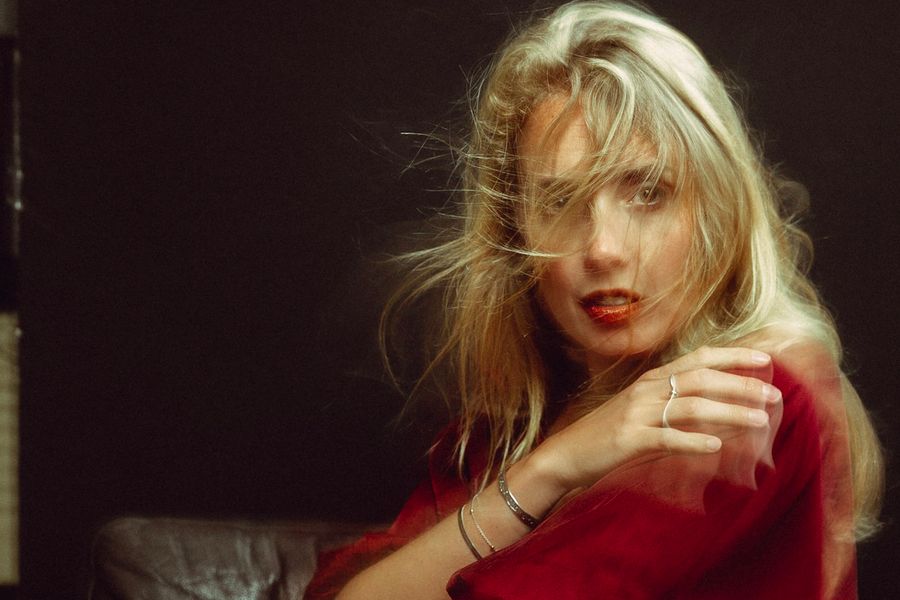 Woman with long blonde hair and a floral dress looks back while holding a transparent umbrella at a crosswalk, with warm café lighting in the background.