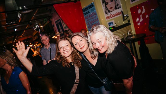 Three women pose happily together during Disco Royale at de Doelen Studio. They smile, stand close, and one waves at the camera. The colorful background with posters and lighting adds to the energetic atmosphere.