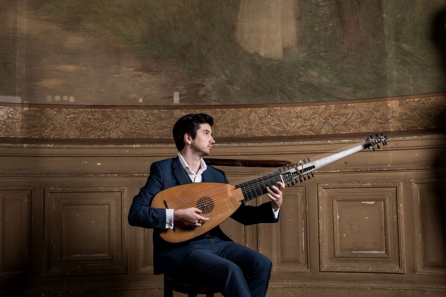 Een man in een donkerblauw pak zit op een houten stoel in een rijkelijk versierde, klassieke ruimte tussen twee stenen pilaren met beeldhouwwerken. Hij speelt een luit en kijkt geconcentreerd naar zijn instrument. De ruimte ademt historische grandeur en akoestische intimiteit.