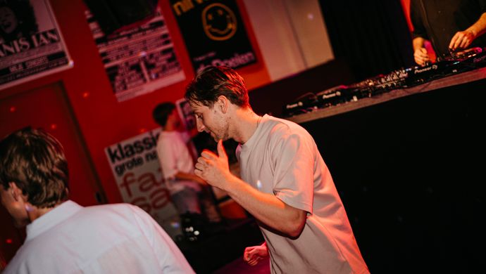 A young man dances during Disco Royale at de Doelen Studio. He stands close to the stage, surrounded by posters and red lighting, with other guests in the background.