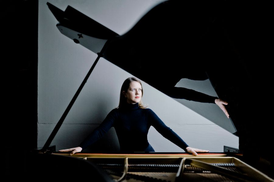 Een vrouw in zwarte kleding zit achter een vleugel in een donkere ruimte. De vleugeldeksel werpt een opvallende schaduw, waardoor er een dramatisch contrast ontstaat tussen licht en donker. De vrouw kijkt geconcentreerd opzij, met haar handen rustend op de rand van het instrument.