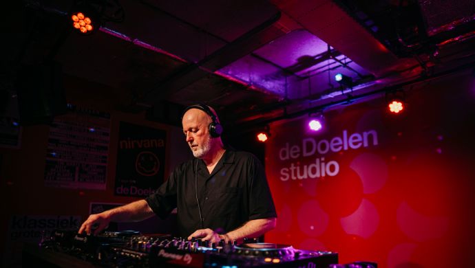 A DJ is focused while playing music during Disco Royale at de Doelen Studio. Wearing headphones, he stands behind a mixing console, surrounded by warm purple and red lighting. On the wall behind him, the logo of de Doelen Studio is visible.