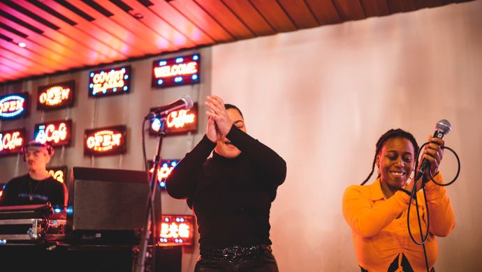 Twee artiesten staan op een podium bij een optreden: de ene persoon in zwarte kleding zingt of spreekt in een microfoon, terwijl de andere persoon in een felgele top enthousiast zingt met een microfoon in de hand. Op de achtergrond is een wand te zien met meerdere schermen of lichtpanelen, wat een levendige sfeer creëert.