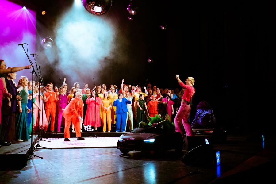The Space Choir tijdens een liveoptreden: een groot koor in kleurrijke outfits zingt onder leiding van een dirigent op een spectaculair verlicht podium.