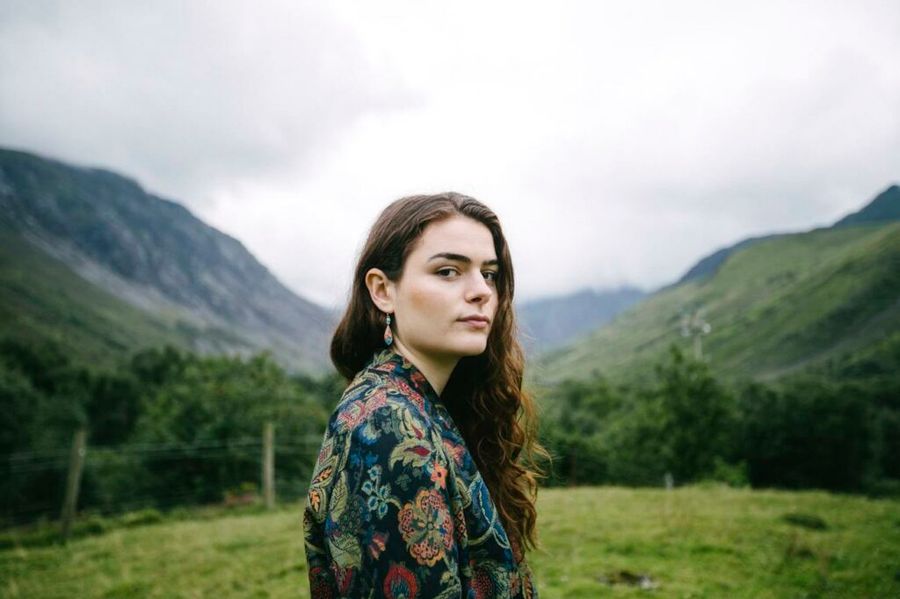 Vrouw met lang bruin haar staat in een groen berglandschap met mistige bergen op de achtergrond. Ze kijkt rustig in de camera, gekleed in een jas met bloemmotief.