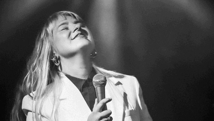 Zangeres in een wit jasje staat onder een spotlight, glimlachend met gesloten ogen en een microfoon in de hand, terwijl het licht haar van boven verlicht.