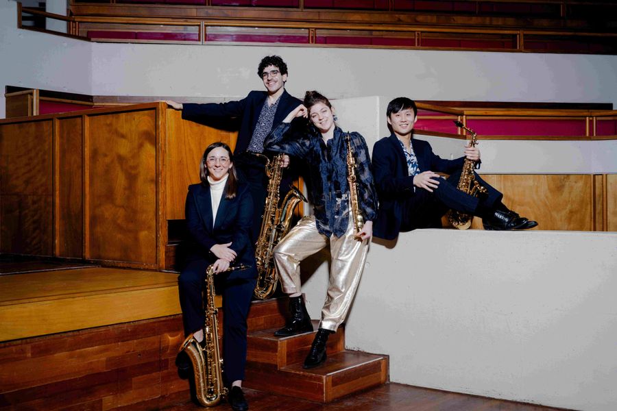 Vier jonge mannen in pak poseren met saxofoons in een modern houten interieur met witte en rode accenten; twee staan op de trap, de andere twee zitten op een verhoging.