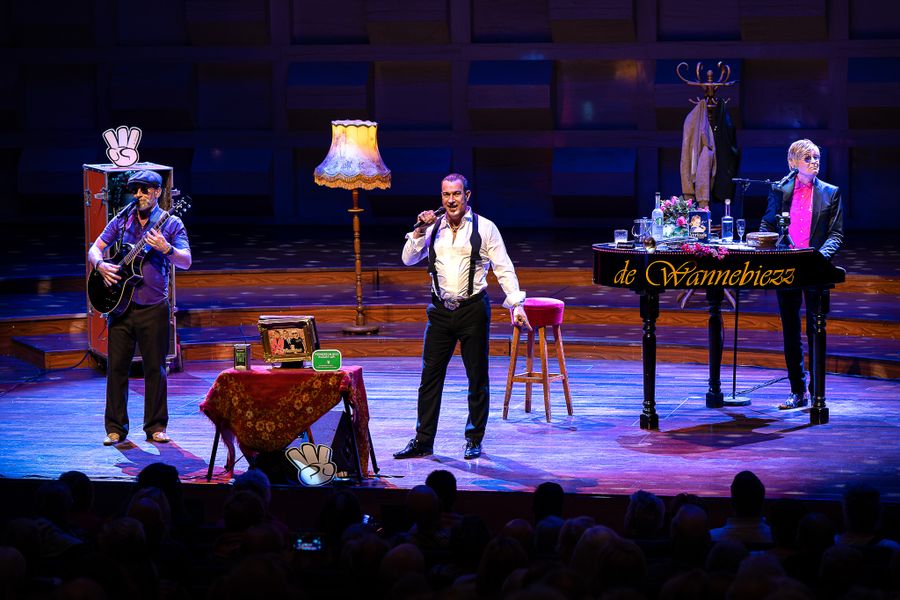 Vier artiesten op een kleurrijk verlicht podium tijdens een theatervoorstelling. In het midden staat een zanger in wit overhemd met bretels, links een muzikant met een accordeon, rechts een pianist, en er staan verschillende huiselijke attributen zoals een eettafel, lampen en een servieskast.