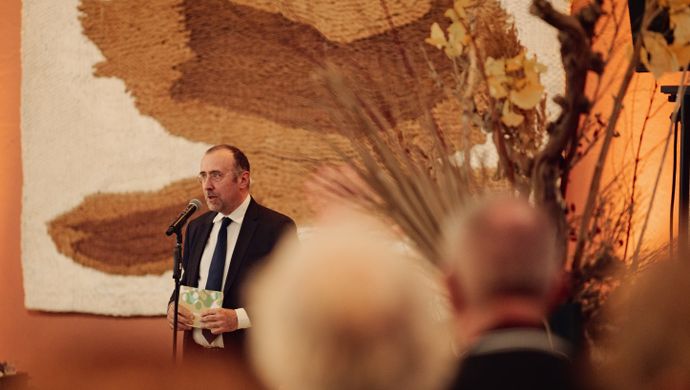 Man in pak met blauwe stropdas en bril kijkt het publiek aan en spreekt in een microfoon