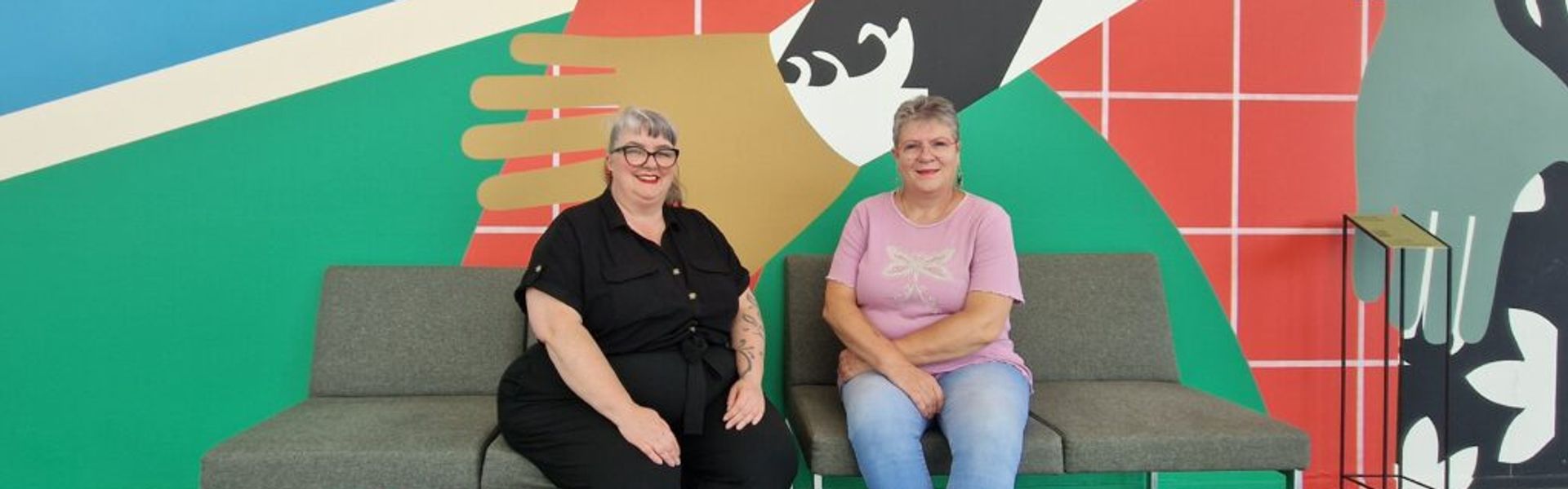 Two women smiling while seated on a bench in front of a colorful wall with graphic shapes and a stylized handshake in the background.