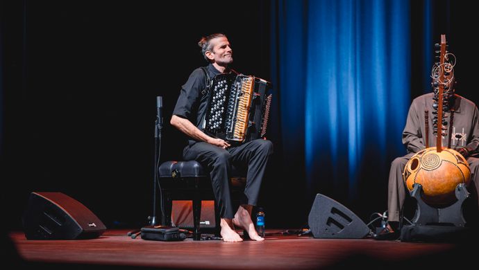 Een accordeonspeler zit ontspannen op het podium en speelt geconcentreerd in warm, sfeervol blauw licht.